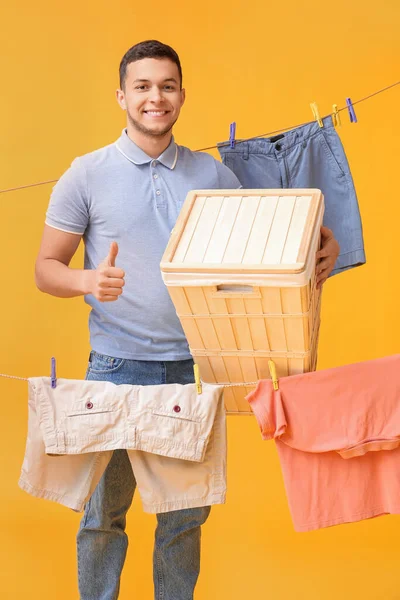 黄色の背景に親指アップを示すバスケット ぶら下げ洗濯やプラスチック製の服を着た若い男 — ストック写真