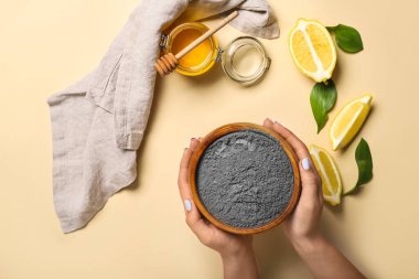 Yüz maskesi için bir kase aktif karbon tozu, renk arkaplanda limon ve bal olan kadın elleri, yakın plan.