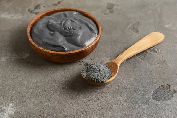 Bowl of activated carbon facial mask and spoon with powder on grunge background