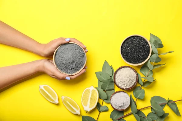 Sarı arka planda aktif karbon yüz maskesi, limon ve okaliptüs dalları için malzeme dolu kadın elleri.