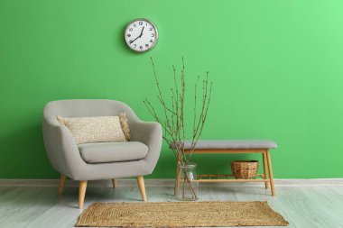 Koltuk, vazo, ağaç dalları, bank ve saat yeşil duvarda asılı.