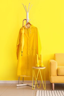 Yağmurluklu raflar, masa üzerinde çizmeler ve sarı duvarın yanında koltuk.