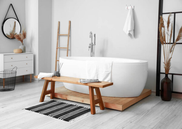 Interior of light room with white bathtub and wooden bench
