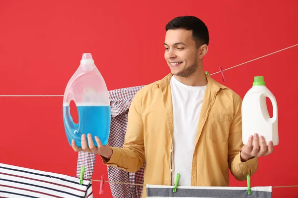 洗剤のボトルを持つ若い男 赤い背景に洗濯や衣類をぶら下げ — ストック写真