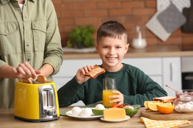 Kızarmış ekmek dilimi ile küçük bir çocuk, bir bardak meyve suyu ve annesi mutfakta.