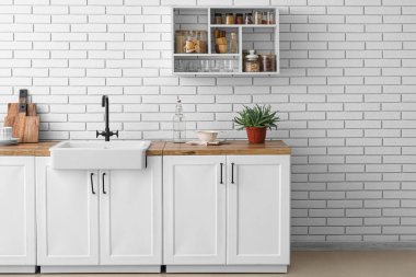 Lavabolu tezgahlar, mutfak gereçleri ve beyaz tuğla duvarda asılı raf ünitesi.