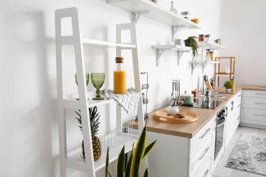 Mutfaktaki beyaz duvarın yanında gözlüklü ve meyve suyu şişeli raf ünitesi.