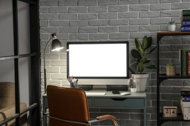 Karanlık odada gri tuğla duvarın yanında bilgisayar, cep telefonu ve parlayan lamba olan işyeri