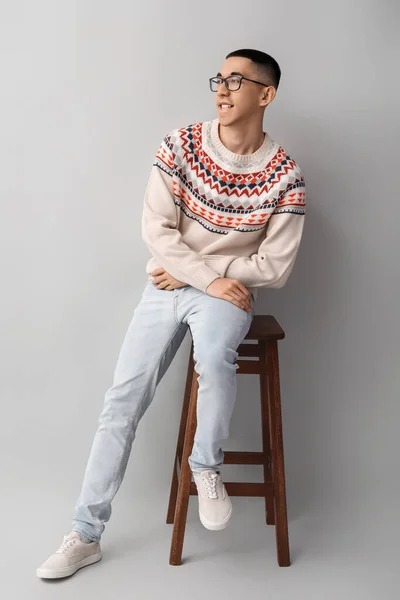 身穿针织毛衣的英俊年轻亚洲男子坐在灰色背景的椅子上 — 图库照片