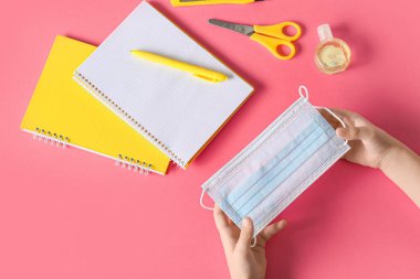 Tıbbi maskeli çocuk elleri, bir şişe dezenfektan ve pembe arka planda kırtasiye malzemesi.