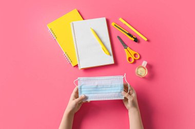 Tıbbi maskeli çocuk elleri, bir şişe dezenfektan ve pembe arka planda kırtasiye malzemesi.