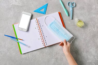 Sağlık maskesi, cep telefonu, dezenfektan ve arka planda kırtasiye malzemesi olan çocuk eli