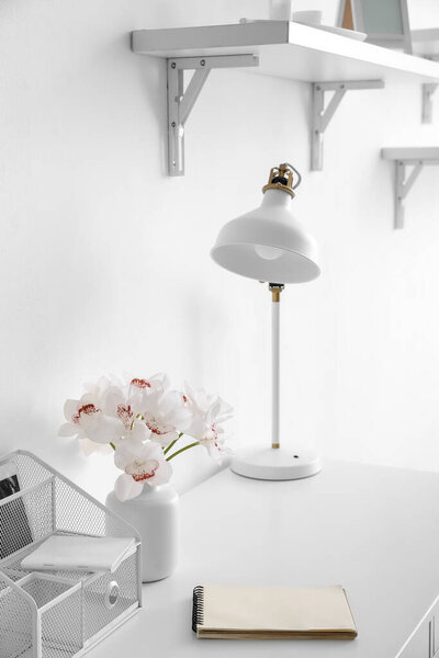 Рабочее место с современной лампой, ноутбуком и вазой с цветами возле белой стены