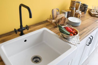 Beyaz lavabo, sebzeli süzgeç ve mutfak gereçleri sarı duvarın yanında.