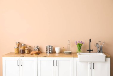 Lavabolu beyaz tezgahlar, vazodaki laleler ve bej duvarın yanındaki mutfak aletleri.