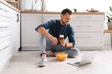 Bilgisayarlı yakışıklı adam bira içiyor ve mutfakta cips yiyor.