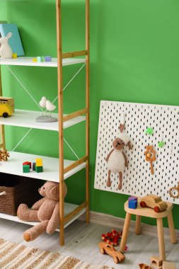 Yeşil duvarın yanındaki taburede oyuncakları ve tahtası olan raf ünitesi.