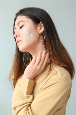 Parlak makyajlı zarif Asyalı kadın ve açık arkaplanda boyuna dokunan şık mücevherler.