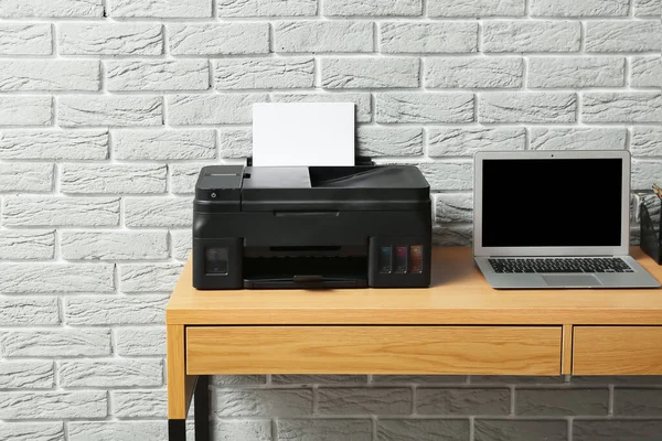 Workplace with modern laptop and printer near grey brick wall