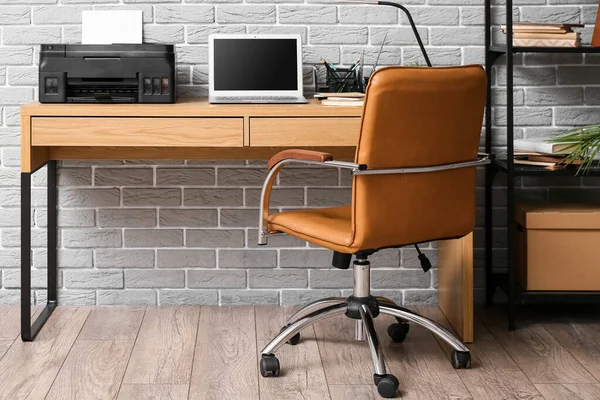 Workplace with modern laptop and printer near grey brick wall in office