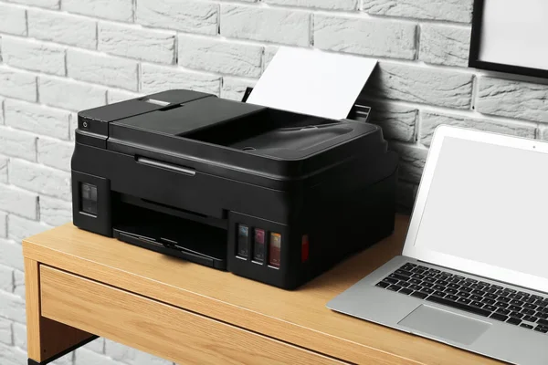 Modern printer and laptop on table near grey brick wall