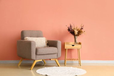 Ahşap masada kuru kamışlı vazo ve pembe duvarın yanında yumuşak koltuk.