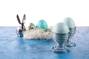 Beyaz arka planda boyanmış Paskalya yumurtaları ve tavşan heykelciği olan bardaklar.