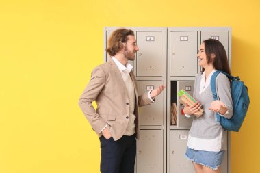 Bayan öğrenci ve öğretmeni sarı arka planda dolabın yanında.