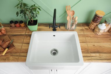 Yeşil duvarın yanındaki tezgahlarda beyaz lavabo, yiyecek ve mutfak aletleri var.