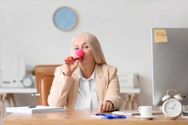 Funny mature woman with clown nose and party whistle in office. April fools day celebration