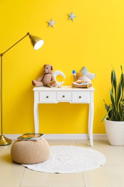 Bebek kıyafetleri, oyuncaklar, lamba ve sarı duvarın yanında saksı.