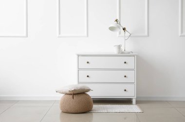 Beyaz duvarın yanında yastıklı ve çekmeceli bej bir puf.