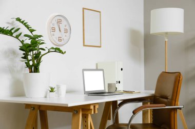 Ofis içi aydınlatma duvarının yanındaki modern dizüstü bilgisayarı, fincanı ve bitkisi olan işyeri