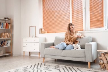 Bir fincan çayla genç bir kadın ve evdeki gri koltukta oturan Jack Russel Terrier.