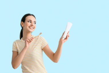 Mavi arka planda diş macunu ve fırçasıyla gülümseyen genç kadın.