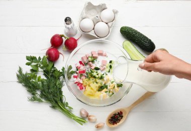Beyaz ahşap arka planda Okroshka için malzemelerle kefir döken kadın.