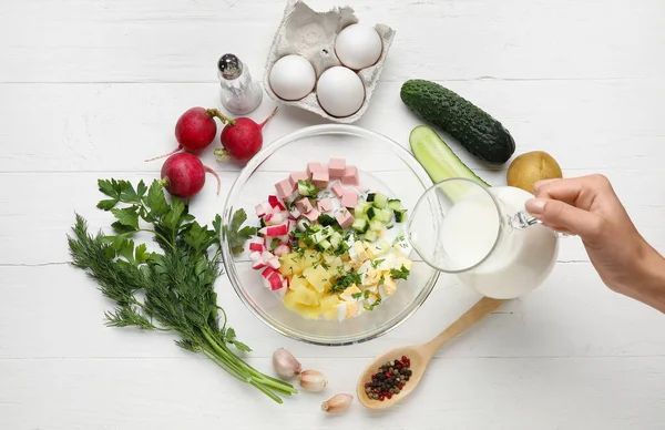 Beyaz ahşap arka planda Okroshka için malzemelerle kefir döken kadın.