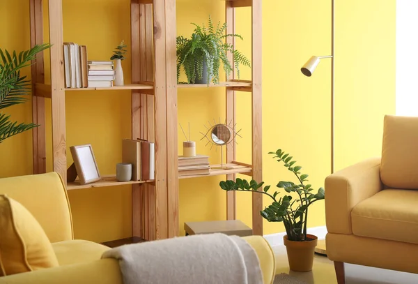 Oturma odasındaki sarı duvarın yanında dekoru ve çiçekleri olan ahşap kitaplık.