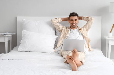 Yatak odasında dizüstü bilgisayar kullanan yalınayak genç bir adam.
