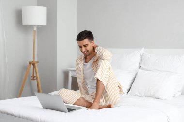 Yatak odasında dizüstü bilgisayar kullanan yalınayak genç bir adam.
