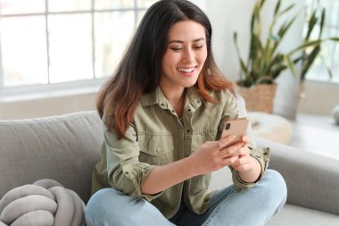 Cep telefonlu güzel bir Asyalı kadın evdeki koltukta oturuyor.