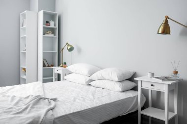 Gri duvarın yanında yumuşak çarşafları, komodinleri, lambaları ve raf ünitesi olan rahat bir yatak.