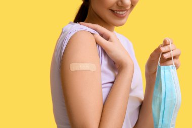 Sarı arka planda uygulamalı tıbbi bandı ve maskesi olan genç bir kadın, yakın plan.