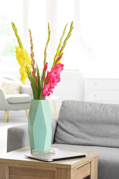 Vase with fresh gladiolus flowers and laptop on table in room