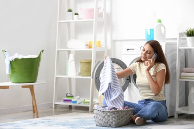 Çamaşırları yıkanmış güzel bir ev hanımı evde çamaşır makinesinin yanında cep telefonuyla konuşuyor.