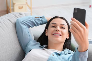 Diş teli takan güzel bir kadın evdeki kanepede selfie çekiyor.