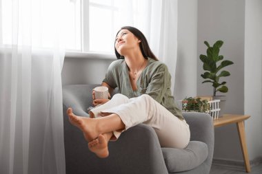 Genç Asyalı çıplak ayaklı bir kadın çay fincanı ile evdeki koltukta oturuyor.