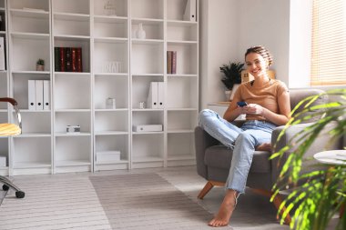 Evdeki koltukta cep telefonu kullanan yalınayak genç bir kadın.