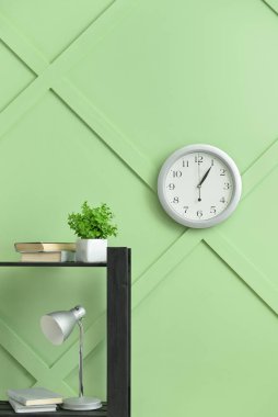 Kitaplı masa, ev bitkisi, lamba ve odanın duvarında asılı modern saat.
