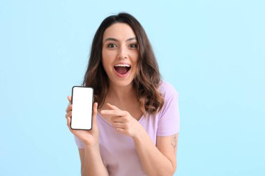Mavi arka planda cep telefonu olan heyecanlı genç bir kadın.